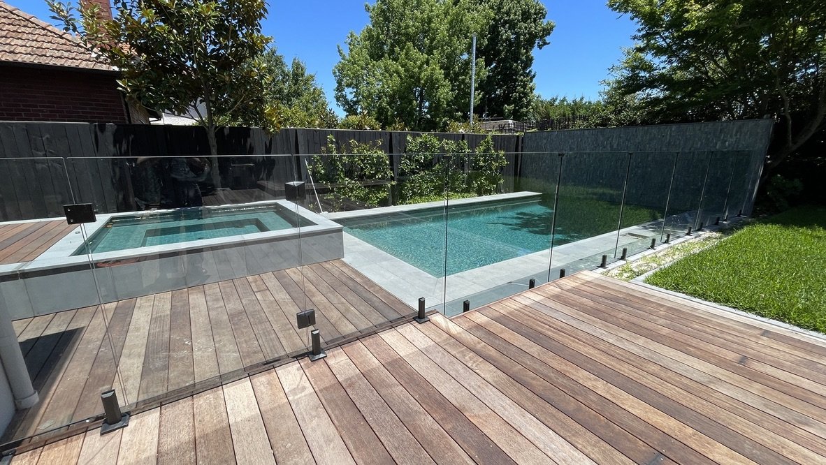 The pool garden in full — timber deck and frameless glass fencing giving onto the pool and spa in soft green, lawn and dark timber boundary