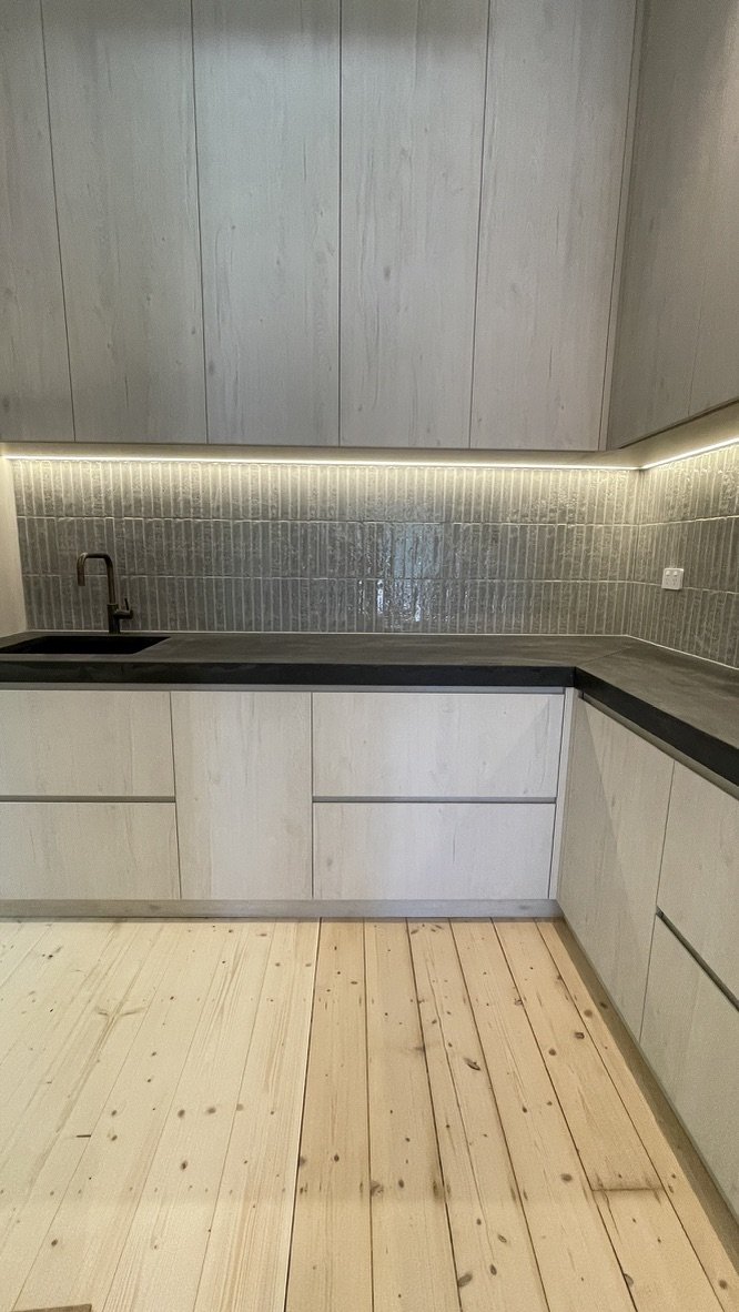 The laundry renewed — grey-washed joinery, dark benchtop and a fluted tile splashback lit from beneath the overhead cabinets