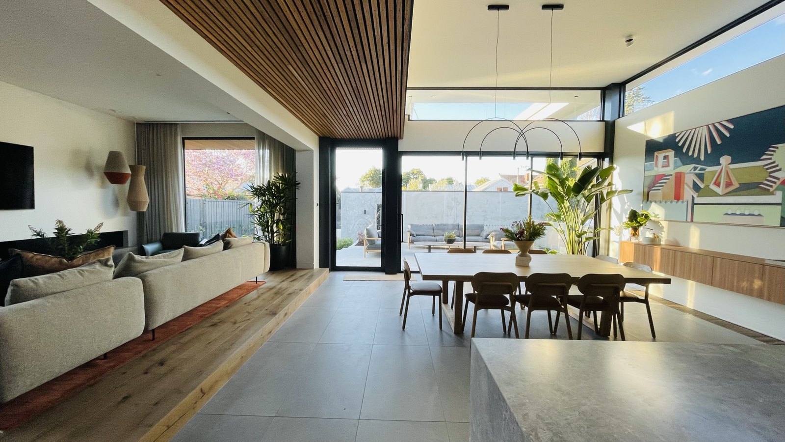 The new rear volume — battened timber ceiling, highline glazing, dining beneath arc pendants, the garden beyond