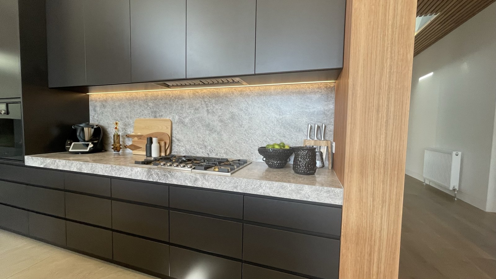 The kitchen — matte black joinery, grey stone benchtop and splashback lit from beneath the overheads, timber column