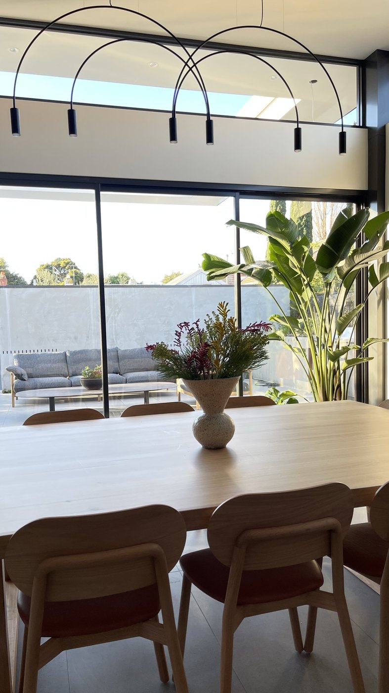 The outdoor lounge seen through the glass from the dining table — grey outdoor sofas and timber, an Amy Wale vase of natives in the foreground