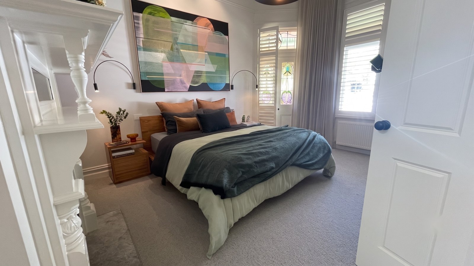 The main bedroom in full — the white-painted original mantel at left, the bed beneath the Ferwerda, stained glass and shuttered windows beyond
