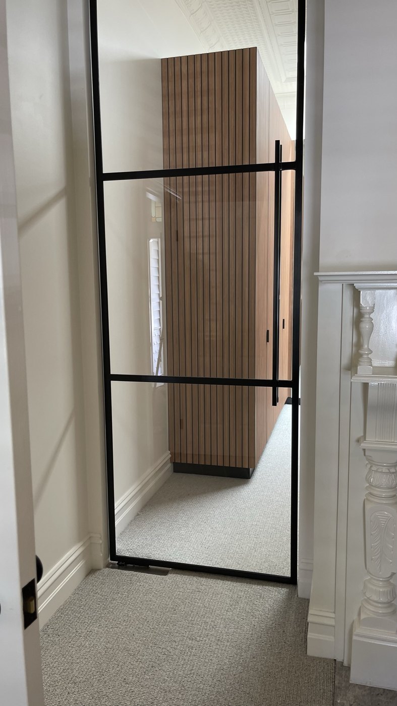 A black steel-framed glass door opening to the walk-in robe, battened timber joinery beyond, original mantel at right