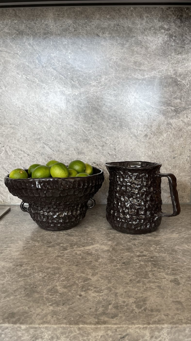 Two dark hand-textured Amy Wale vessels on the stone bench — a bowl of limes and a jug