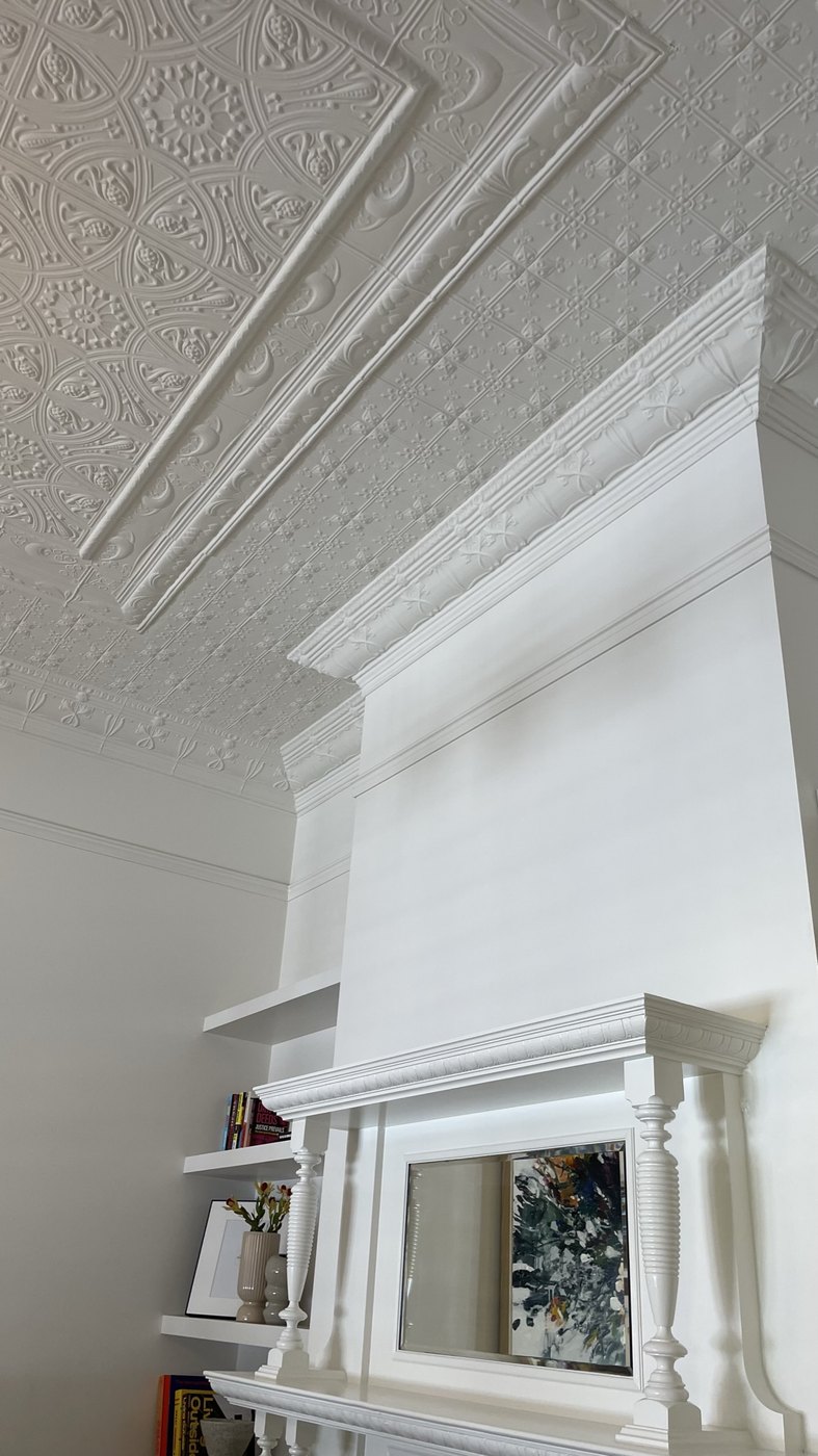 The overmantel restored in white beneath the retained pressed-metal ceiling and stepped cornices