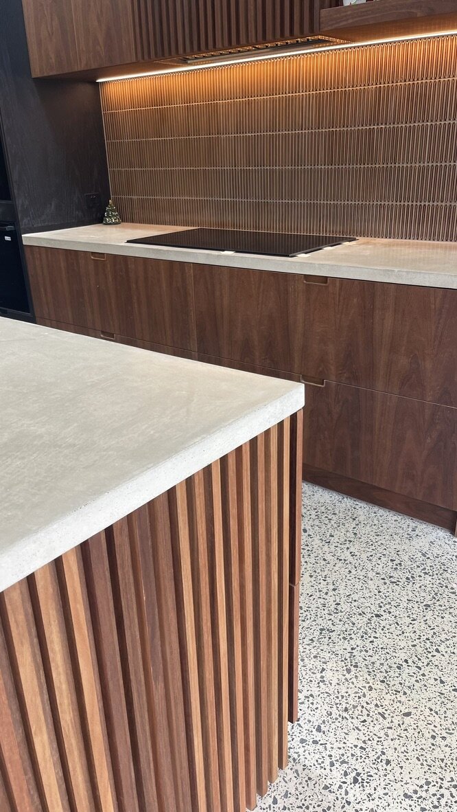 The island — ribbed spotted gum beneath a thick polished concrete benchtop, the fluted caramel splashback glowing behind