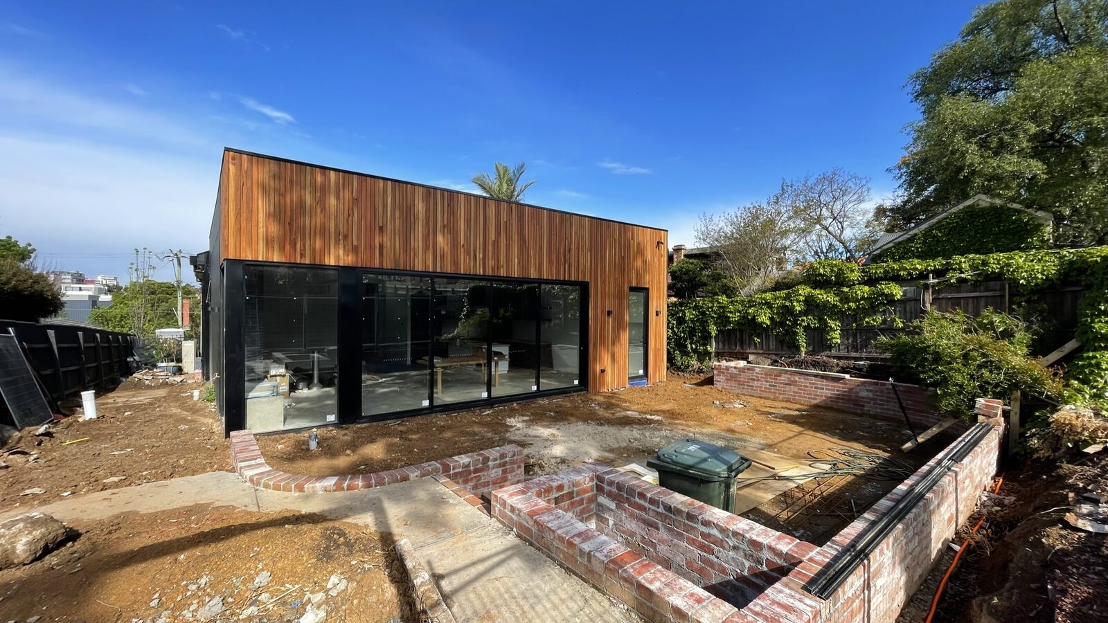 The rear elevation nearing completion — spotted gum and black cladding above recycled brick garden beds taking shape